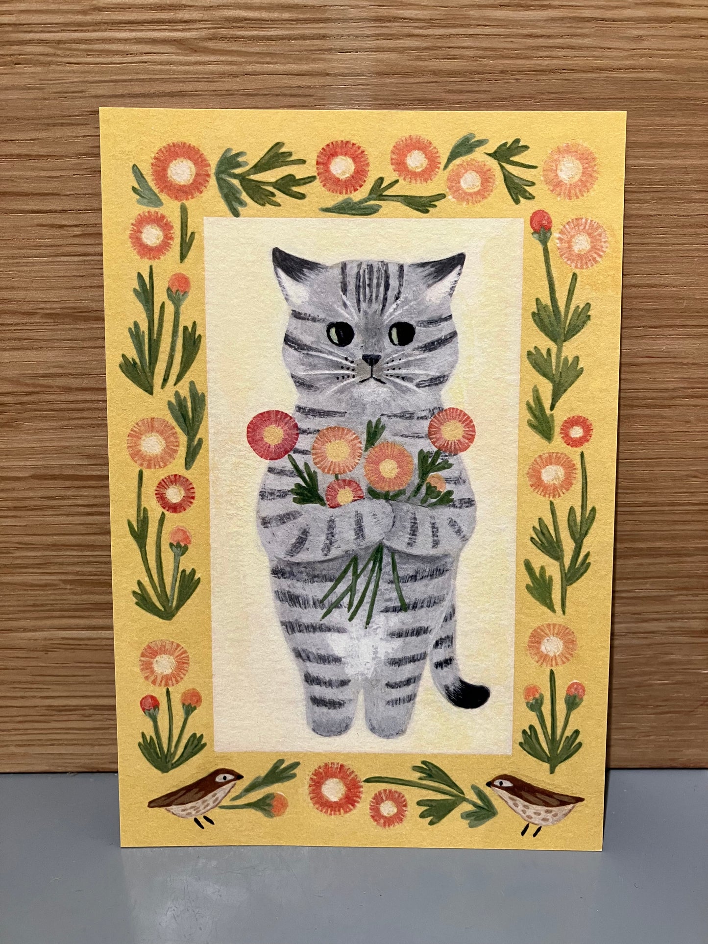 Japansk kort gul baggrund, med gråstribet kat der holder blomster